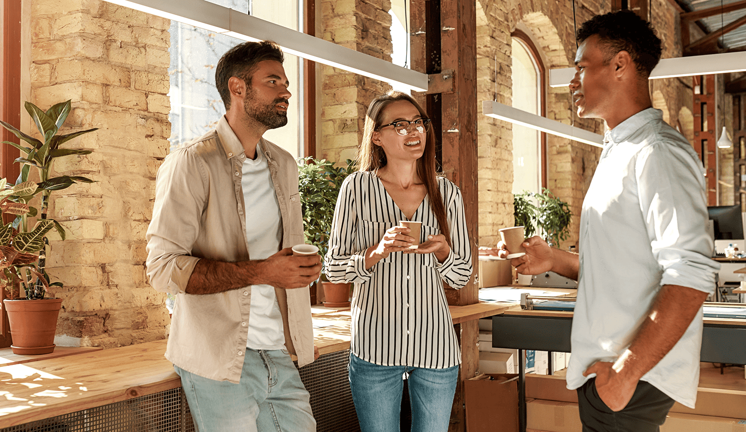 Three colleagues stand in a bright, modern office, talking casually while holding coffee cups in a friendly and collaborative workplace atmosphere. The space has large windows, exposed brick walls, wooden details, plants, and warm natural light.