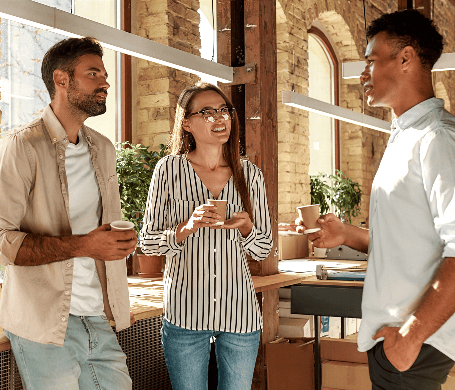 Three colleagues stand in a bright, modern office, talking casually while holding coffee cups in a friendly and collaborative workplace atmosphere. The space has large windows, exposed brick walls, wooden details, plants, and warm natural light.