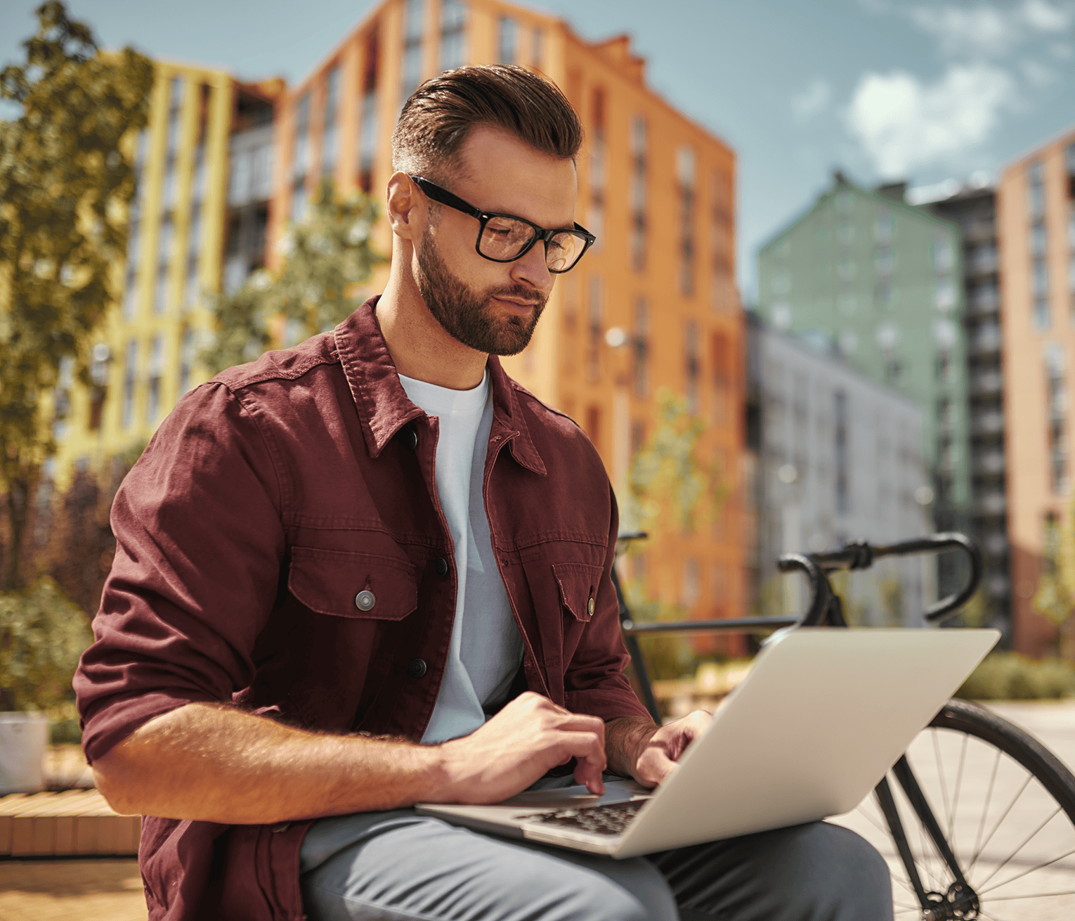 A man working on a laptop outdoors next to his bicycle, with colorful apartment buildings in the background.