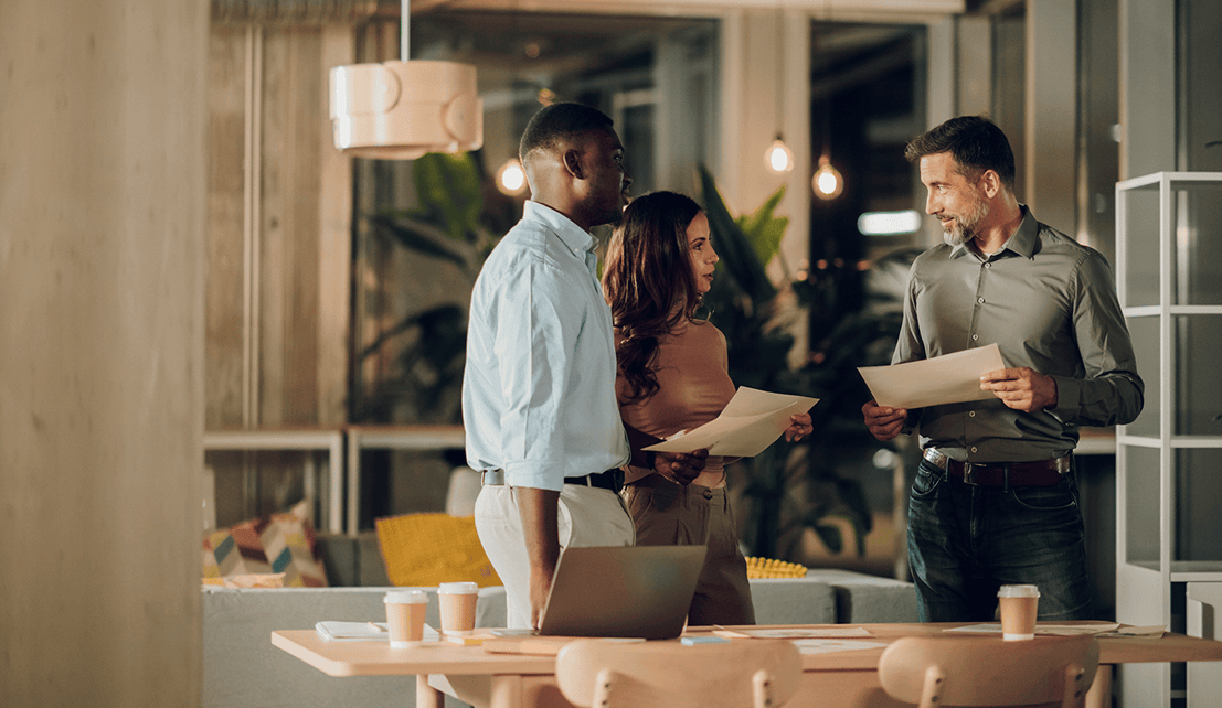 Three colleagues standing around a table in a modern office, reviewing printed documents beside an open laptop and coffee cups.
