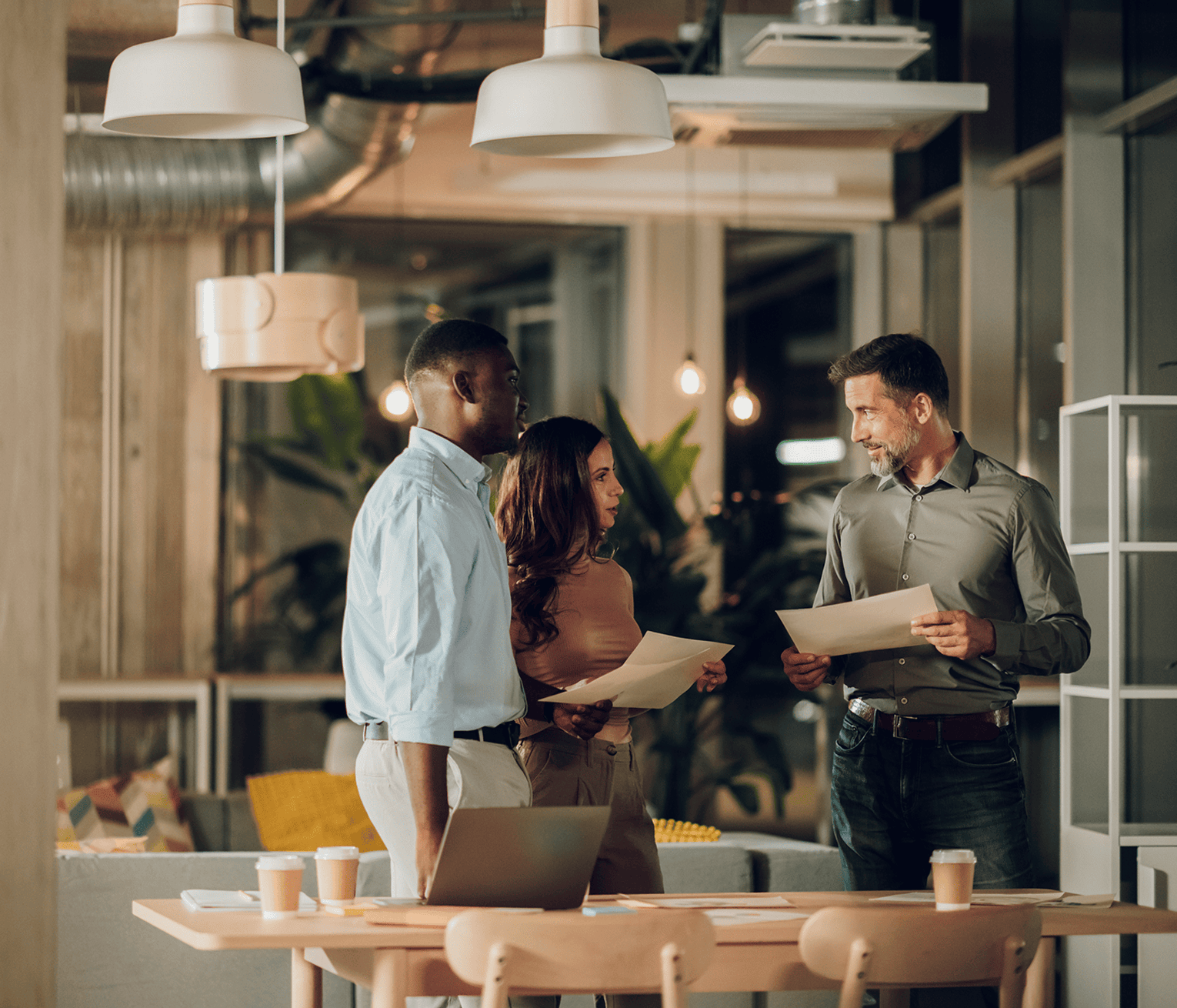 Three colleagues standing around a table in a modern office, reviewing printed documents beside an open laptop and coffee cups.