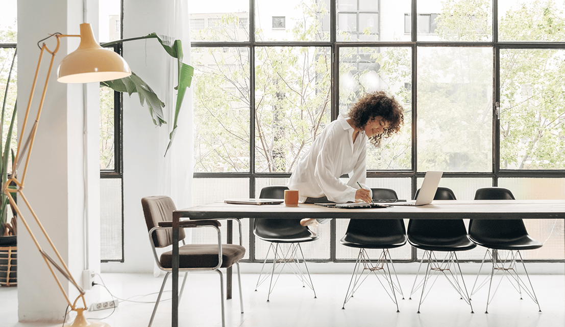 Professional working at a long table with a laptop in a bright, modern office with floor-to-ceiling windows.