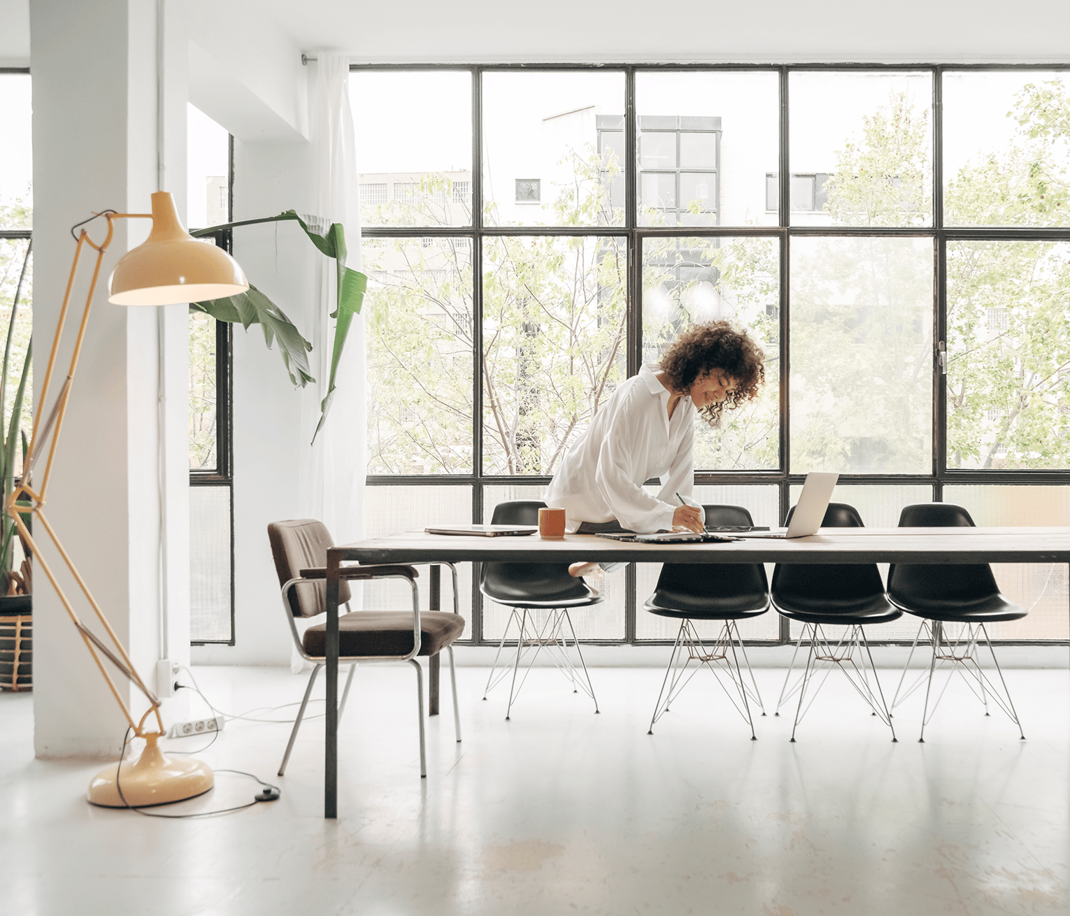 Professional working at a long table with a laptop in a bright, modern office with floor-to-ceiling windows.