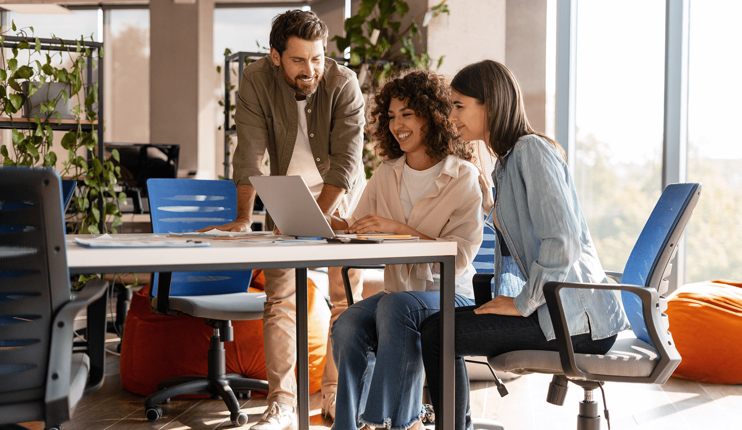 Three smiling colleagues gathered around a laptop in a bright modern office, collaborating together at a shared desk.