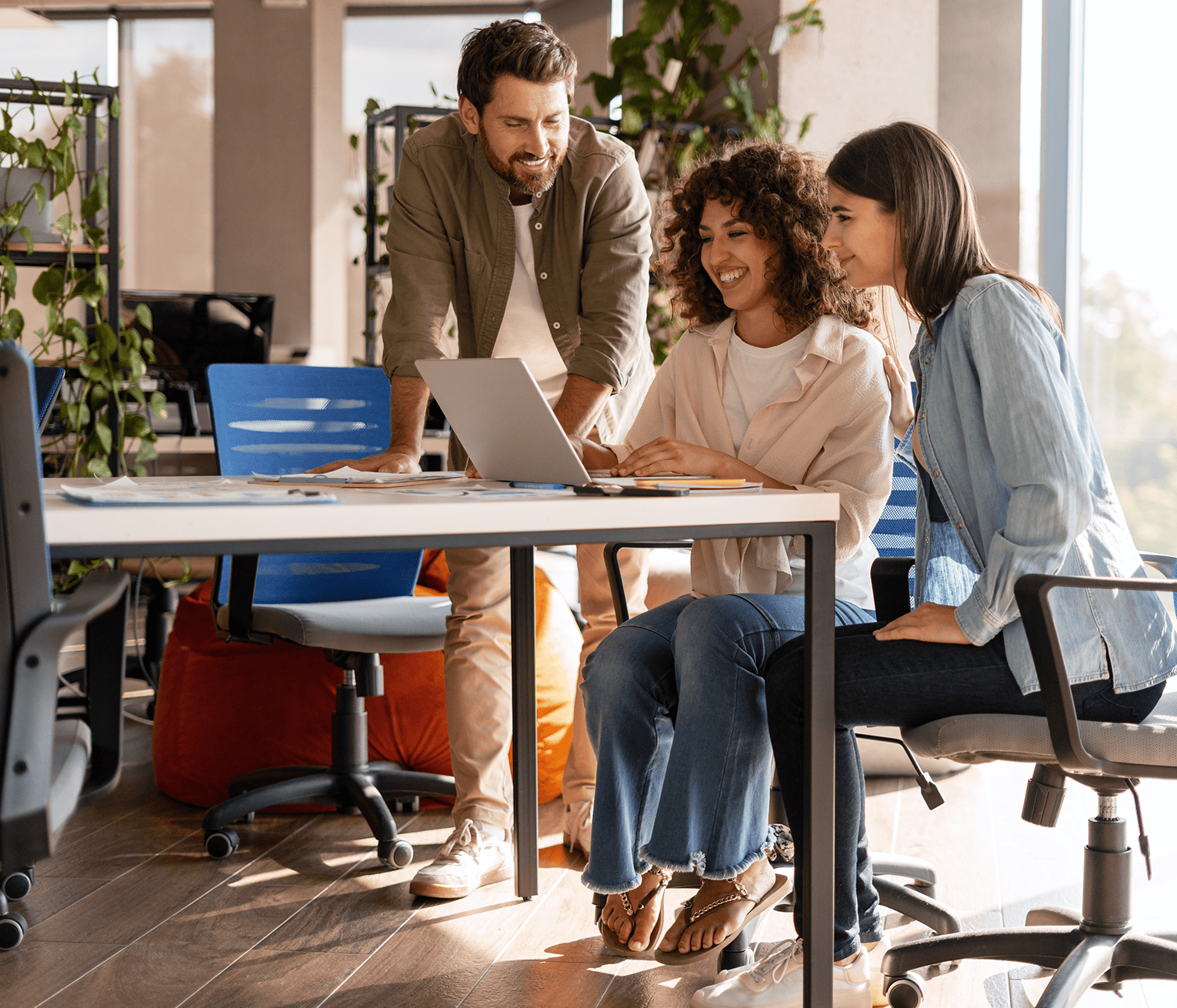 Three smiling colleagues gathered around a laptop in a bright modern office, collaborating together at a shared desk.