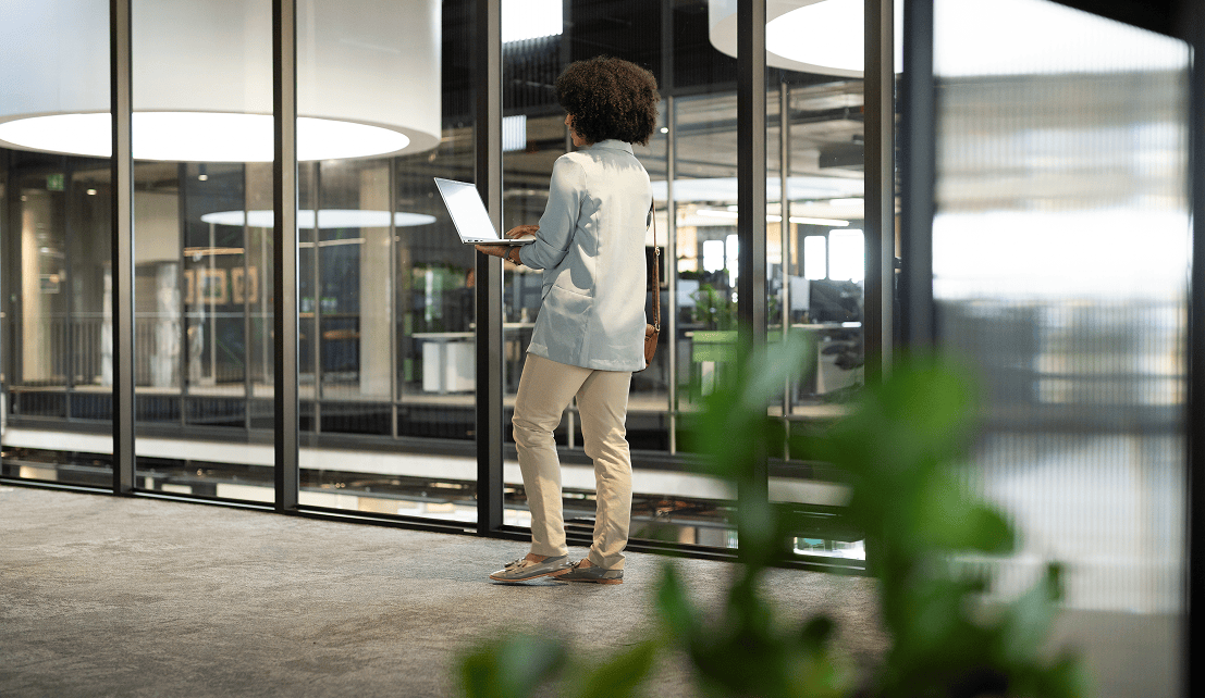 Female employee holding a laptop while standing by large windows overlooking a modern office.
