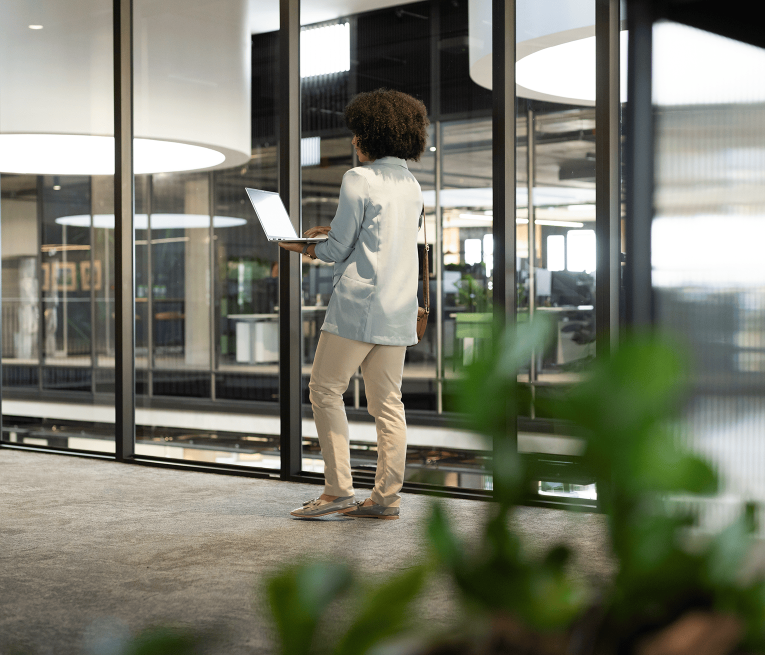 Female employee holding a laptop while standing by large windows overlooking a modern office.