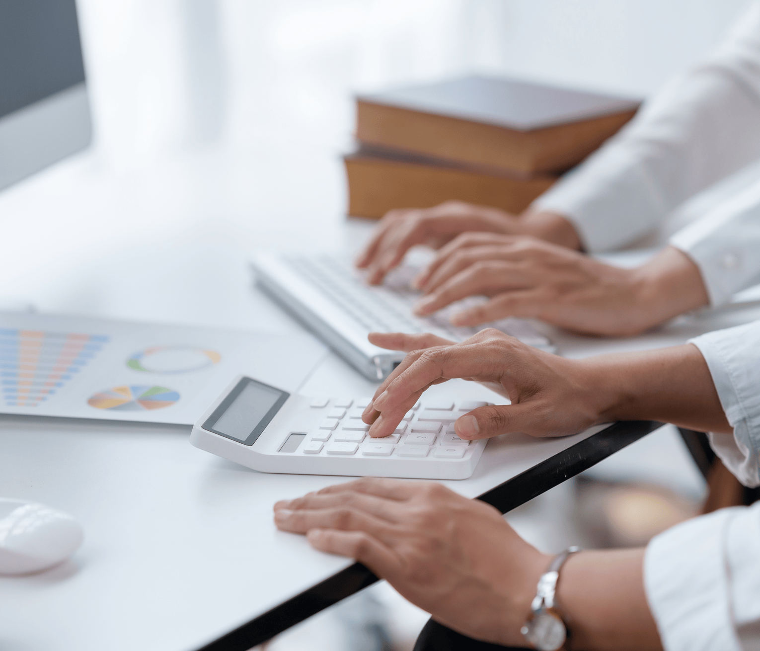 Close-up of two people’s hands wearing white shirts working at a desk in front of a computer, one using a calculator while the other types on a keyboard, with printed charts and graphs nearby.