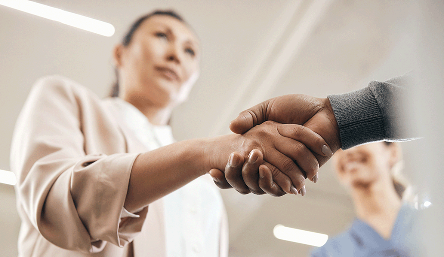 Two professionals shaking hands in an office setting, with a smiling colleague standing in the background.