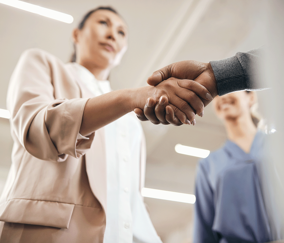 Two professionals shaking hands in an office setting, with a smiling colleague standing in the background.