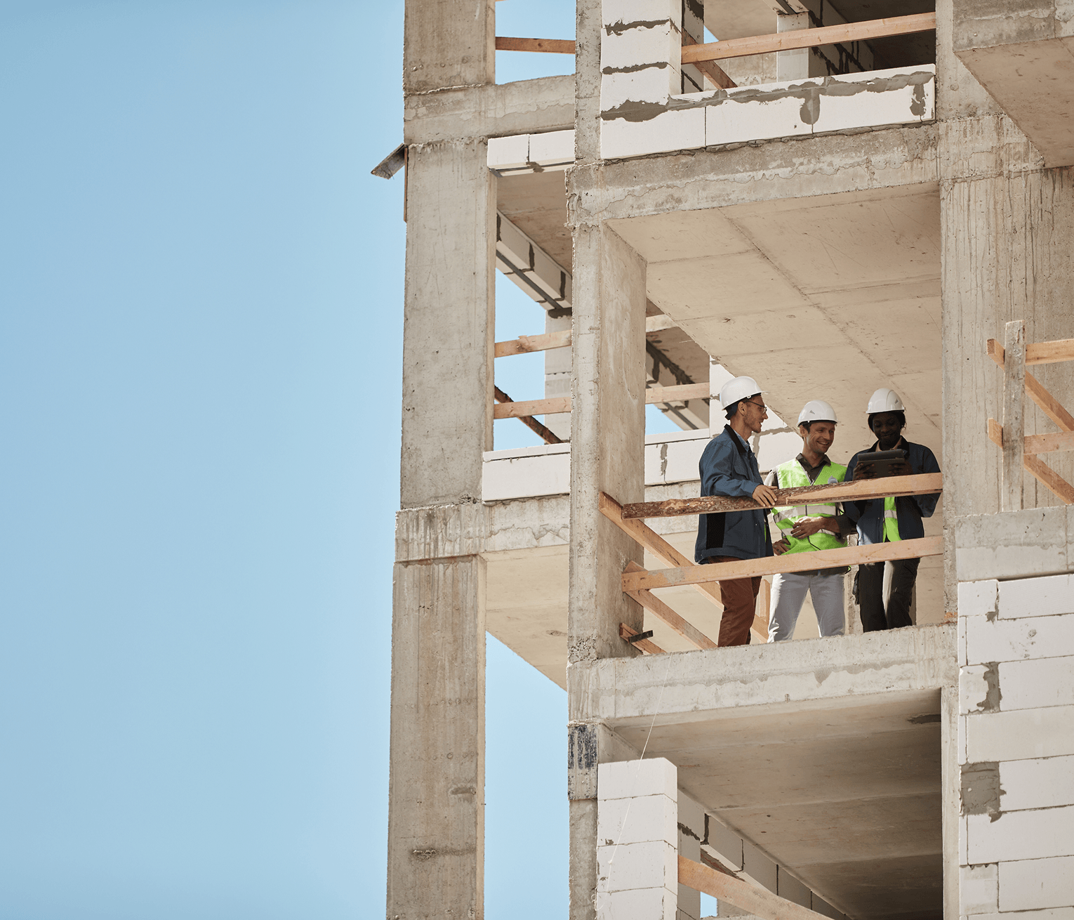 Three construction professionals wearing safety helmets stand on the upper floor of a building under construction, reviewing project plans together.