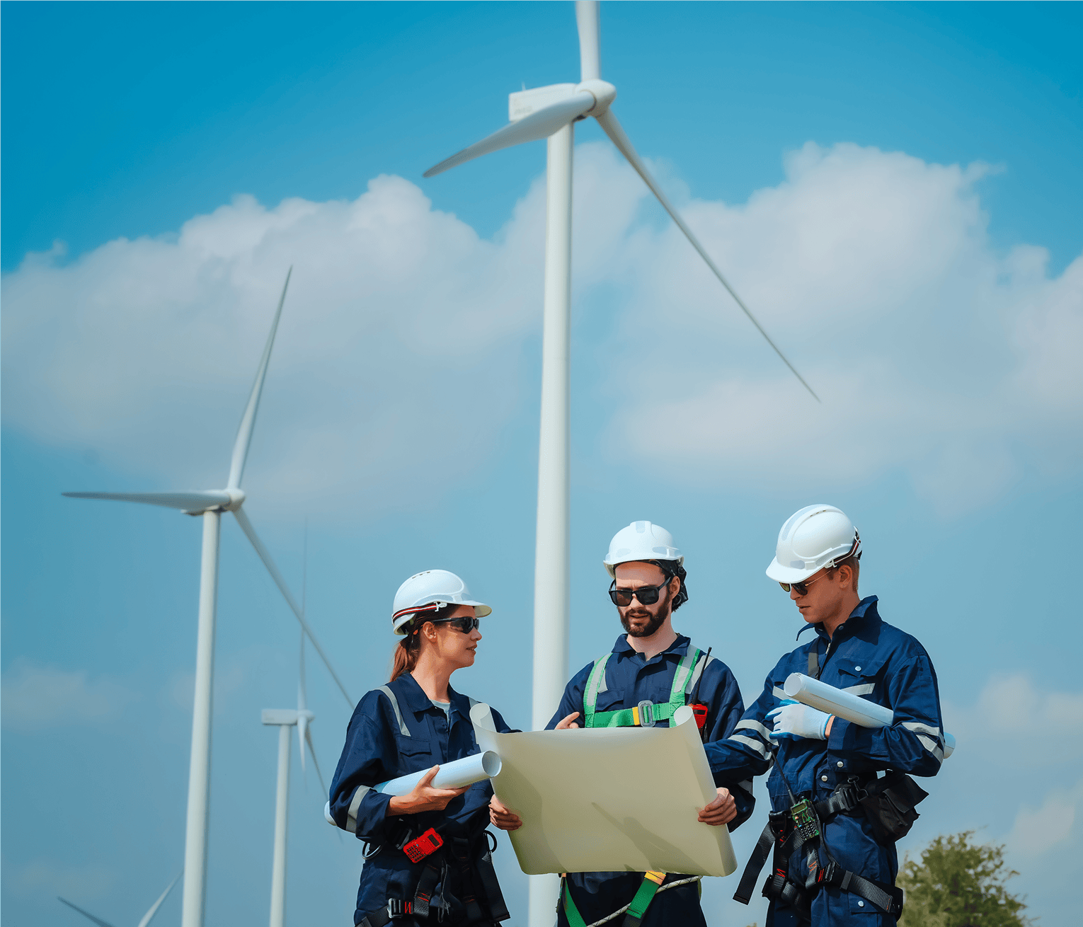 Three wind farm engineers wearing safety helmets review technical plans together in front of wind turbines under a blue sky.