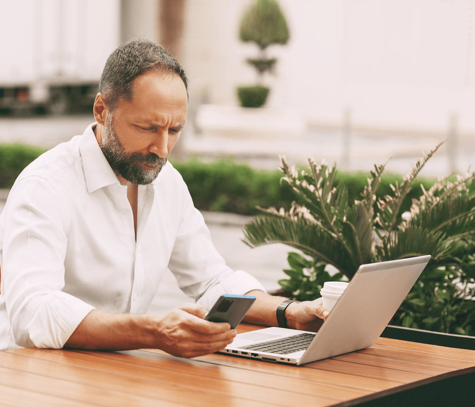 Man multitasking on a laptop while checking his phone while sitting at an outdoor café table.