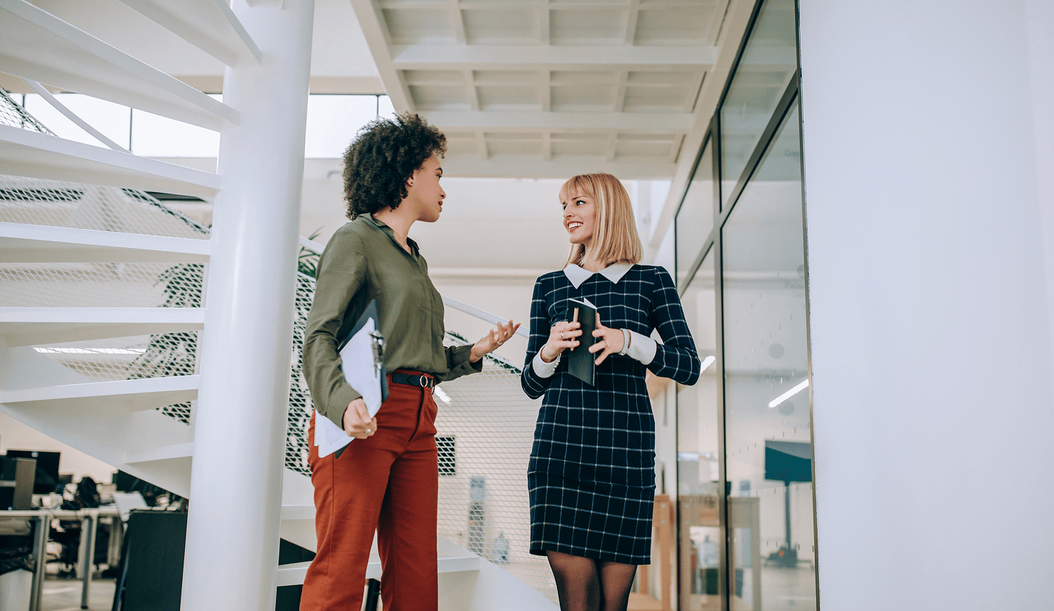 Two female professionals in conversation in a bright, modern office corridor.