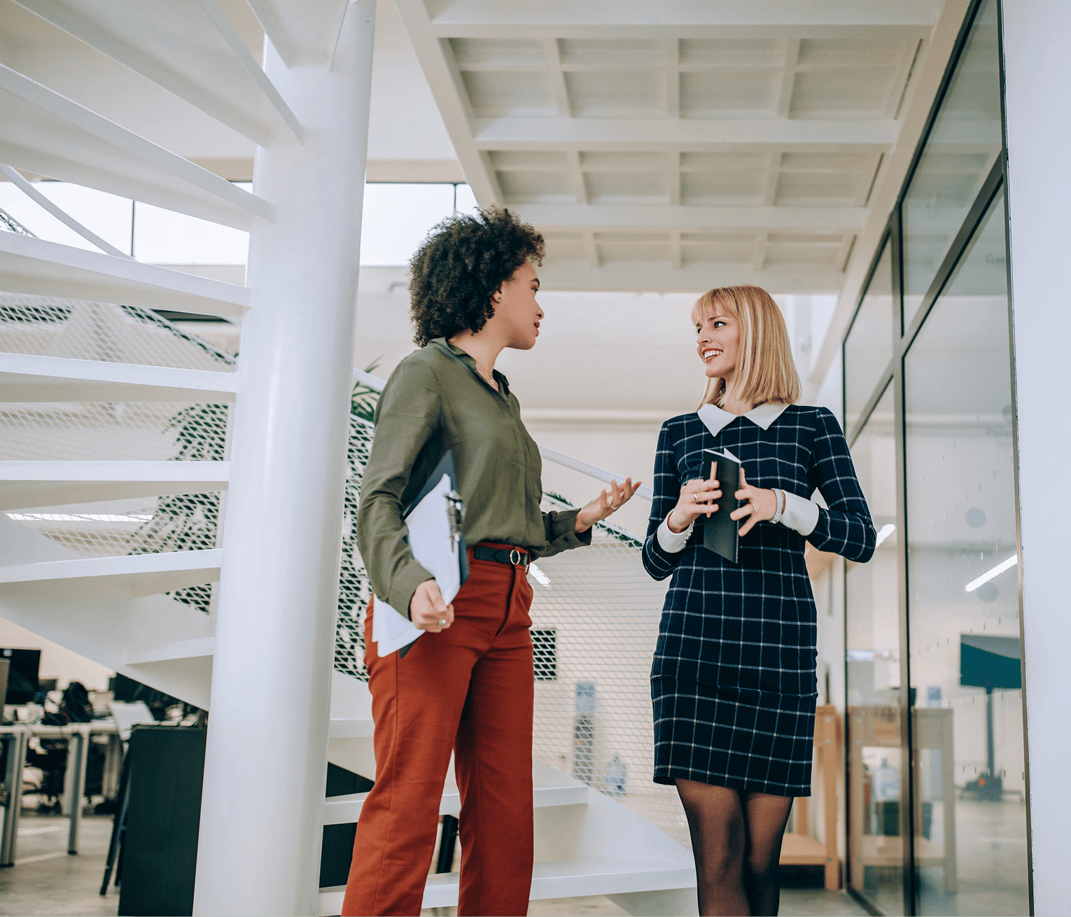Two female professionals in conversation in a bright, modern office corridor.