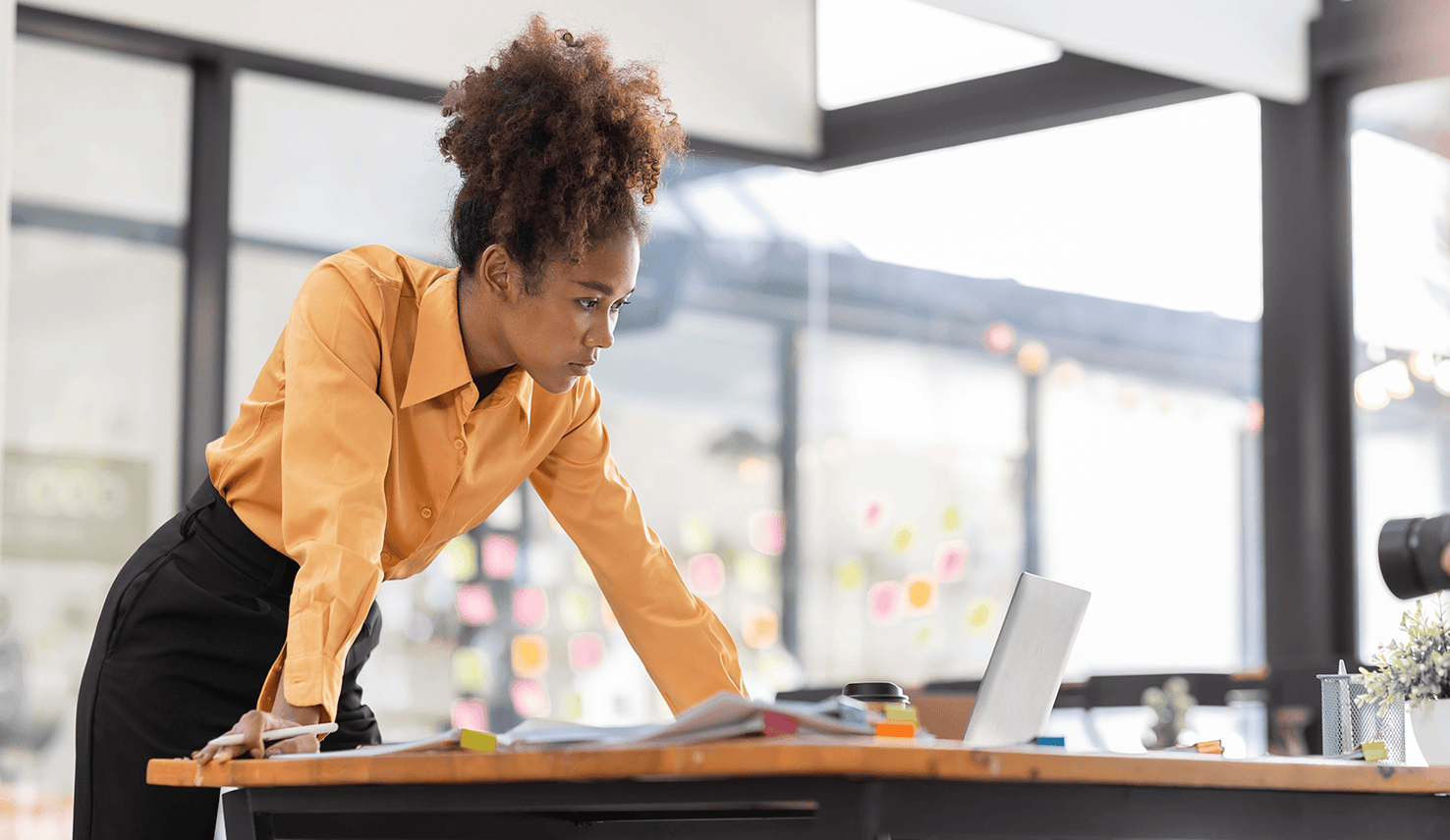 Professional leaning over a desk, reviewing documents and working on a laptop in a bright office environment.