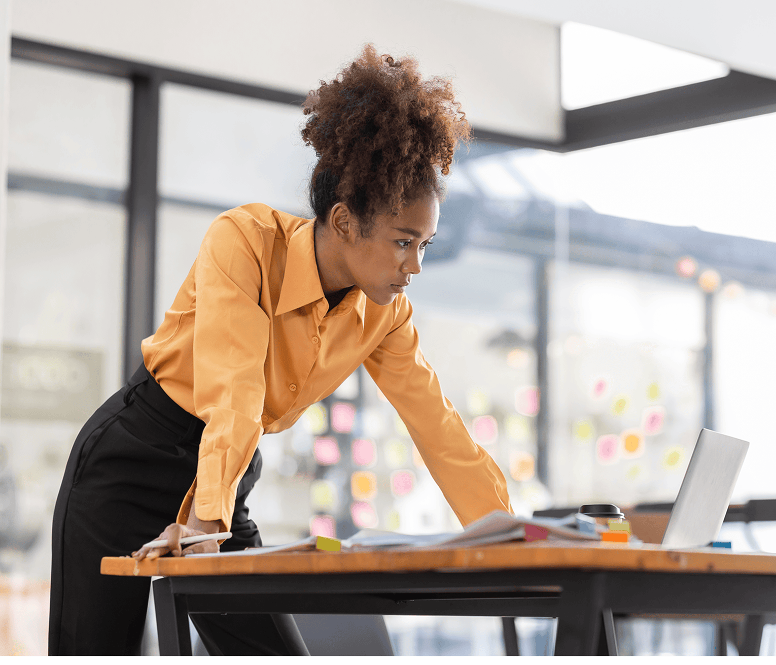 Professional leaning over a desk, reviewing documents and working on a laptop in a bright office environment.