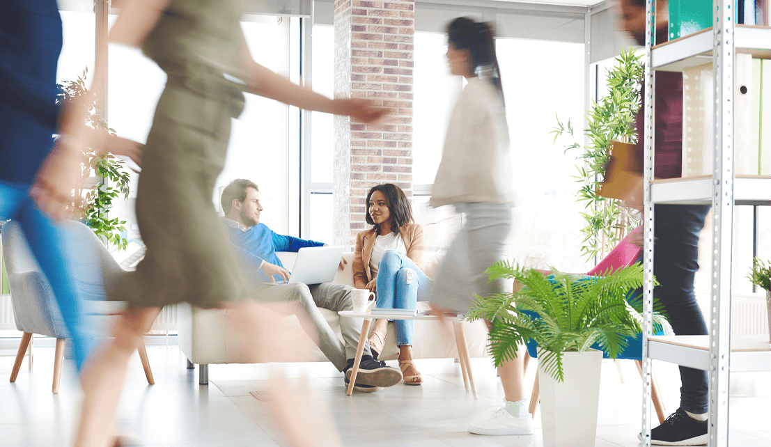 Two colleagues talking on a sofa in the background as employees move through a busy office.