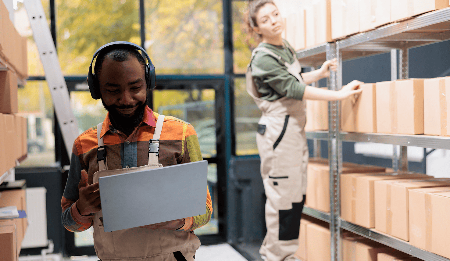 Employee reviewing information on a laptop in a warehouse while a colleague organizes inventory on storage shelves.